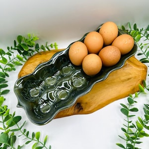 May include: A dark green ceramic egg tray with a wavy, elongated shape featuring twelve circular indentations. Six brown chicken eggs are nestled into one end of the tray, which is resting on a natural, light-colored wooden base.