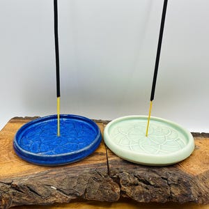 May include: Two ceramic incense holders. One is a vibrant blue, the other a soft green. Both have a circular design and a central hole for incense sticks. The holders sit on a rustic wooden surface.