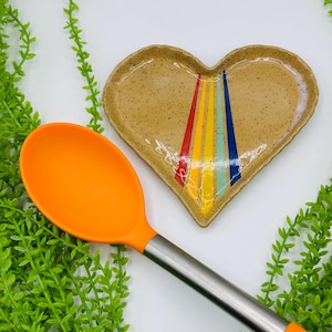 May include: A heart-shaped ceramic dish with a speckled tan finish and a central rainbow stripe pattern in red, orange, yellow, green, and blue. Beside the dish is a kitchen spoon with a bright orange silicone head and a metallic silver handle.