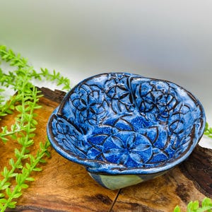 May include: A handmade, blue ceramic bowl with a floral pattern. The bowl has a unique, organic shape and a dark brown rim. The interior features a detailed, embossed floral design. The bowl is displayed on a piece of wood with green foliage.