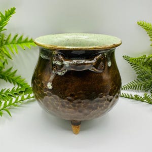 May include: A round, three-legged ceramic cauldron with a dark brown, textured exterior and a smooth, light green interior. The pot features a horizontal handle near the rim with white speckled glaze accents dripping down the sides.