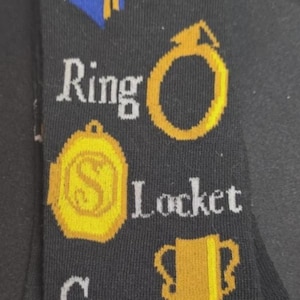 May include: Black socks with gold and white graphics featuring the words "Diary", "Ring", "Locket", and "Cup". The socks also have a gold ring, a gold locket, and a gold cup design. The top of the socks have the words "Harry Potter".