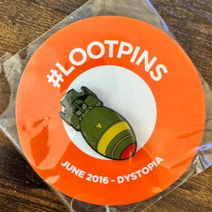 May include: A pin featuring a cartoon bomb design in green, yellow, and red, set against a white circle on an orange background. The pin is labeled with the text "#LOOTPINS" and "JUNE 2016 - DYSTOPIA".