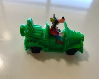 May include: A green toy jeep with a cartoon wolf driver. The jeep has a black steering wheel and black wheels. The wolf is wearing a red shirt and brown pants.