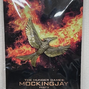 May include: A pin featuring a golden bird with outstretched wings, set against a fiery background. The pin is on a card with the text "THE HUNGER GAMES: MOCKINGJAY PART 2" and "THE FIRE WILL BURN FOREVER ONLY IN CINEMAS".