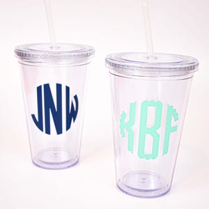 Op de afbeelding: Twee doorzichtige plastic bekers met deksels en rietjes. Eén beker heeft een marineblauw monogram met de letters JNW, de andere een mintgroen monogram met de letters KBF.