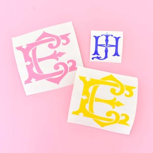 May include: Three adhesive vinyl decals featuring decorative lettering. One large pink letter E, one large yellow letter E, and one smaller blue monogram featuring the letters H and J intertwined in a stylized, ornate font on white backing paper.