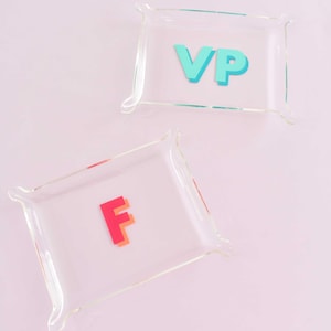 May include: Two clear acrylic trays with rounded corners. One tray has the letters "VP" in turquoise, and the other has the letter "F" in red. The trays are on a light pink background.
