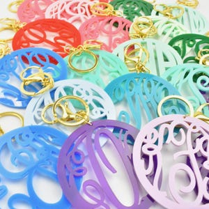 Puede incluir: Una colección de dijes de llavero de acrílico de colores con un diseño de monograma. Los dijes son de varios colores, incluyendo rosa, rojo, verde, azul, morado y blanco. Cada dije tiene un llavero dorado.