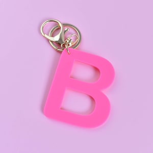 Puede incluir: Un llavero con la letra "B" de color rosa brillante con un cierre y un anillo dorados. La letra tiene un diseño sólido y audaz, sobre un fondo rosa suave y liso.