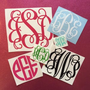 May include: A collection of monogram decals in various colors and sizes. The decals feature elegant, swirling letter designs in pink, white, green, turquoise, and black. The decals are displayed on a glittery pink surface.
