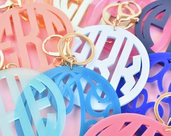 May include: A collection of circular acrylic keychains in various colors including light blue, dark blue, coral, pink, and mint green. Each keychain features a cutout monogram design and is attached to a gold-toned metal lobster clasp ring.