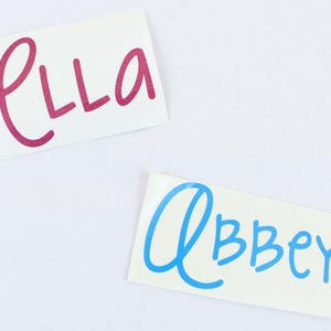 May include: Two rectangular white stickers with names written on them. One sticker has the name "Ella" in pink cursive. The other sticker has the name "Abbey" in blue cursive.