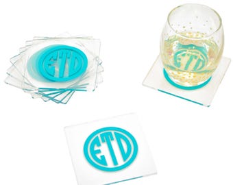 May include: A set of square clear acrylic coasters featuring a circular turquoise monogram with the letters ETD. One coaster is placed individually, one is under a clear glass, and a stack of several coasters is shown to the left of the glass.