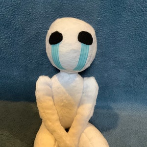 Op de afbeelding: Een witte pluche pop met een ronde kop, zwarte ogen en blauwe traanvormige accenten. De pop heeft een eenvoudig lichaam en armen gekruist voor zich. De pop zit op een blauw getextureerd oppervlak.