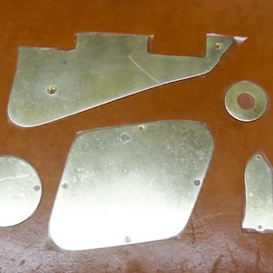 May include: A collection of gold-colored guitar parts, including pickguards and covers, are arranged on a brown surface. The parts vary in shape and size, with screw holes visible on some. The components appear to be new and unused.