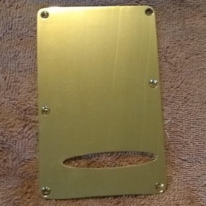 May include: A gold-colored metal guitar pickguard with a rectangular shape and rounded corners. It has a small oval cutout in the center and several small holes around the edges.