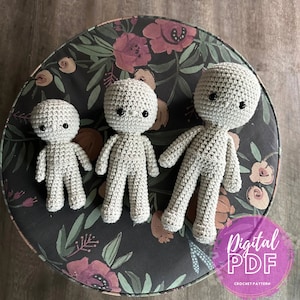 Peut inclure: Trois poupées en crochet de tailles différentes, toutes de couleur gris clair, avec des yeux noirs. Les poupées sont assises sur une surface en tissu floral foncé. Le texte "Modèle de crochet PDF numérique" est visible dans le coin inférieur droit de l'image.