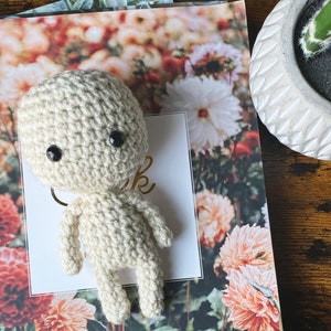 May include: A handmade, cream-coloured crochet doll with black button eyes. The doll is posed on a floral print card that says "thank". A small, white, carved pot with a cactus is in the background. The doll has a simple, minimalist design.