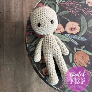Op de afbeelding: Een gehaakte pop met een beige lichaam en zwarte ogen. De pop zit op een oppervlak met een bloemenpatroon. De tekst "Digital PDF Crochet Pattern" is zichtbaar in de rechterbenedenhoek van de afbeelding.