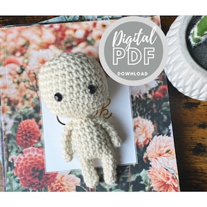 Op de afbeelding: Een gehaakte pop in lichtbeige, met een ronde kop, kleine armen en benen en zwarte knoogogen. De pop ligt op een boek met bloemenpatroon. Een cirkel toont "Digital PDF Download".