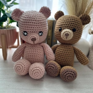 Puede incluir: Dos osos de peluche de ganchillo hechos a mano. Uno es rosa claro, el otro marrón. Ambos tienen ojos de seguridad negros y narices pequeñas y redondas. Los osos están sentados sobre una superficie de color claro, con los brazos y las piernas extendidos.