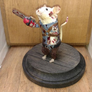 May include: A white mouse wearing a blue and brown outfit with blood splatters, holding a red and black chainsaw. The mouse is standing on a dark wood base.