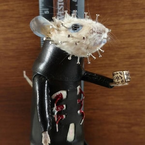 May include: A small, taxidermied mouse wearing a black leather outfit with red accents and a small gold box in its hand. The mouse has a blue eye and is covered in pins.