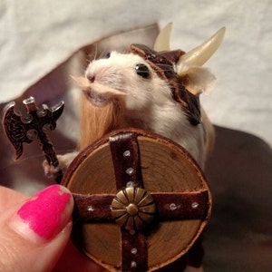 May include: A white mouse dressed as a Viking warrior with a brown leather helmet, a brown beard, a silver axe, and a round wooden shield with a brown leather strap and a brass center piece.