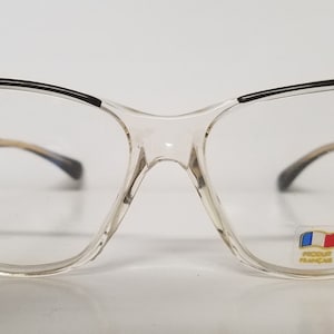 Puede incluir: Gafas de plástico transparente de ojo de gato con monturas negras y detalles dorados. Las gafas tienen una pequeña pegatina de la bandera francesa en la lente derecha con el texto "Made in France".