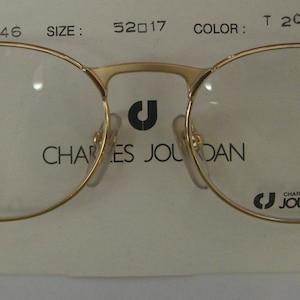Puede incluir: Gafas redondas de color dorado con lentes transparentes. La montura tiene el texto "CHARLES JOURDAN" impreso en la parte delantera. El número de estilo es CJ 146, la talla es 52017 y el color es T 206.