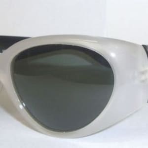 Puede incluir: Gafas de sol blancas y negras con montura de ojo de gato. Las lentes son de color gris oscuro.