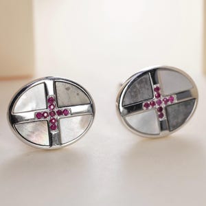 May include: A pair of oval-shaped cufflinks featuring a silver-toned metal frame. Each cufflink is divided into four quadrants containing alternating white mother-of-pearl and grey stone inlays, centered with a cross-shaped cluster of small red rubies.