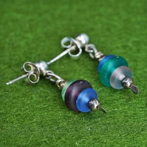 May include: A pair of handcrafted earrings with a post-back closure. Each earring features a silver-toned metal design with a series of colorful, frosted glass beads in shades of blue, green, purple, and clear. The earrings are displayed on a green surface.