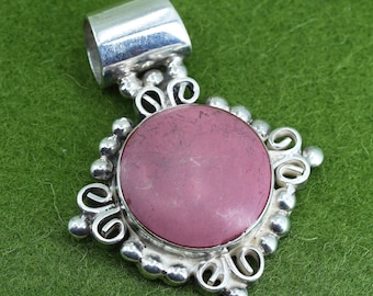 May include: A silver pendant with a large, round, dusty rose-colored stone at the center. The stone is set in a silver frame with decorative swirls and small silver beads. The pendant has a silver loop at the top for a chain.