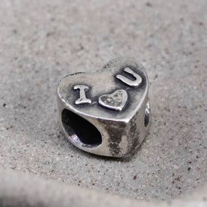 Puede incluir: Un charm plateado en forma de corazón con las letras "I U" y un pequeño símbolo de corazón. El charm tiene un acabado envejecido y dos agujeros para ensartar. Para una pulsera o collar.