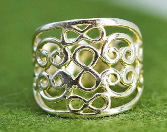 Puede incluir: Anillo plateado con un diseño calado de símbolos de infinito y espirales repetidos. El anillo tiene una banda ancha y un acabado pulido, sobre un fondo verde. El intrincado patrón es visible en esta toma de cerca del anillo.