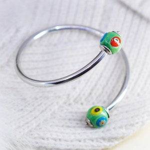 May include: A silver-tone, open-ended bangle bracelet with two green, glass beads. Each bead has a floral design with red, blue, and yellow accents. The bracelet is approximately 2.5 inches in diameter.