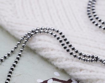 May include: Close-up of two strands of a silver ball chain necklace. The chains are draped over a white, textured surface. The small, round beads of the necklace reflect light, creating a subtle shine. The background is slightly blurred.