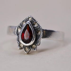 May include: A silver ring featuring a teardrop-shaped, deep red gemstone. The gemstone is set in a decorative, ornate setting with small gold-colored accents. The ring band is a simple, polished silver.