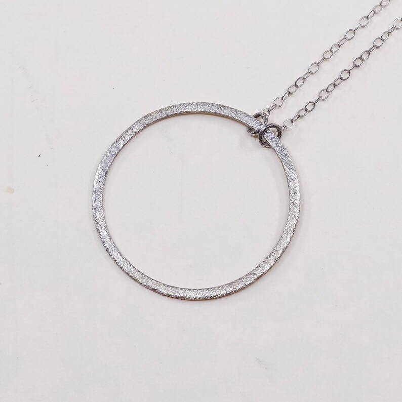 May include: A silver-toned necklace featuring a textured, open circle pendant. The pendant is connected to a delicate chain. The design is simple and elegant, suitable for everyday wear or special occasions.