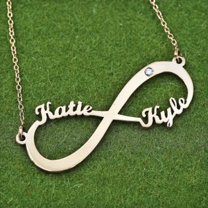 May include: Gold-colored infinity necklace with the names "Katie" and "Kyle" in a cursive font. A small, clear gemstone is set in the infinity symbol. The necklace is displayed on a green surface.