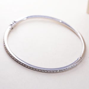 May include: A silver-toned bangle bracelet with a row of small, clear gemstones set along the top. The bracelet has a hinged clasp and a smooth, polished finish. The overall design is simple and elegant.