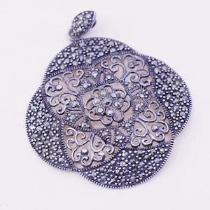 May include: A silver-toned pendant with a floral design. The pendant features a central flower surrounded by intricate filigree and small, sparkling stones. The pendant has a loop at the top for a chain.