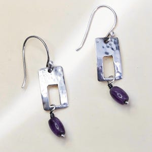 May include: A pair of silver-toned drop earrings featuring a rectangular frame with a hammered texture. Each earring has a small, smooth, oval-shaped purple bead suspended from the bottom of the frame by a thin wire wrap and a curved ear wire hook.