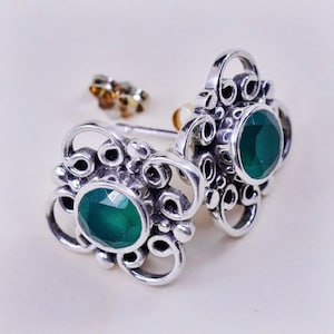 May include: A pair of sterling silver stud earrings with a floral design. Each earring features a faceted, oval-cut, dark green gemstone set in a decorative silver frame. The earrings have a post and butterfly backing.