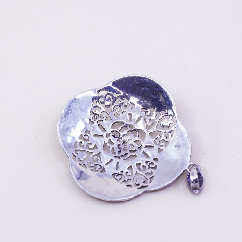 May include: A silver-toned pendant with a floral design. The pendant features a layered, openwork design with a central flower and intricate, swirling patterns. A small loop is attached for hanging.