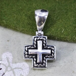 May include: A silver-toned pendant in the shape of a cross. The cross has a detailed design with a rope-like border. The bail is a simple, curved shape. The pendant is likely made of metal and is suitable for a necklace.