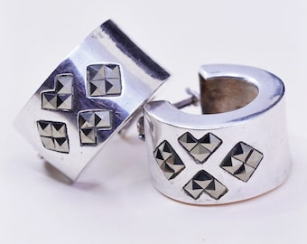 Puede incluir: Un par de pendientes de aro plateados. Cada pendiente presenta cuatro diseños cuadrados con detalles geométricos de color gris oscuro. Los pendientes tienen un acabado pulido y un diseño sencillo y moderno.