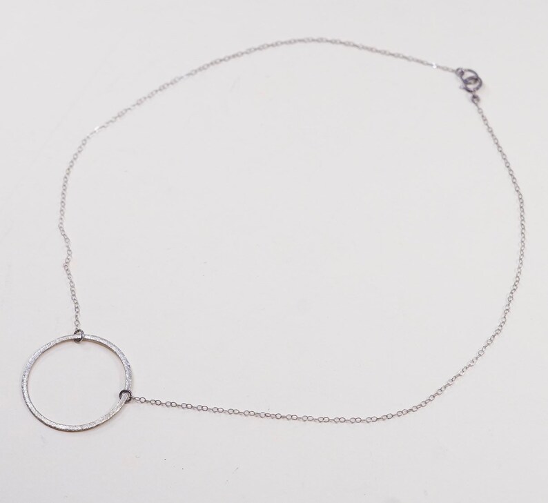 May include: A delicate silver necklace featuring a circular pendant. The pendant is connected to a fine chain that fastens with a small clasp. The necklace is displayed on a white background, highlighting its minimalist design.